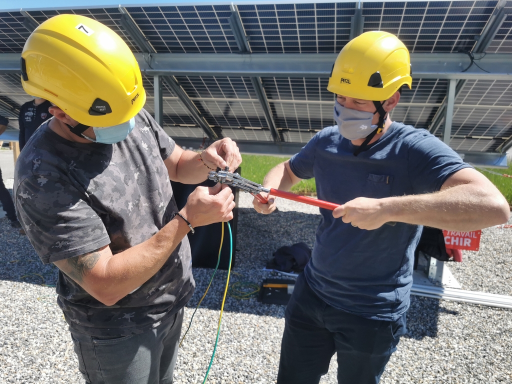 Les stagiaires qualipv et le formateur travaillent ensemble sur de nouvelles compétences au cours de formations professionnalisantes et qualifiantes de courte durée. Ils travaillent sur l'installation de panneaux photovoltaïques. Deux stagiaires sertissent un embout de câble de terre durant la phase pratique d'une formation qualipv photovoltaïque